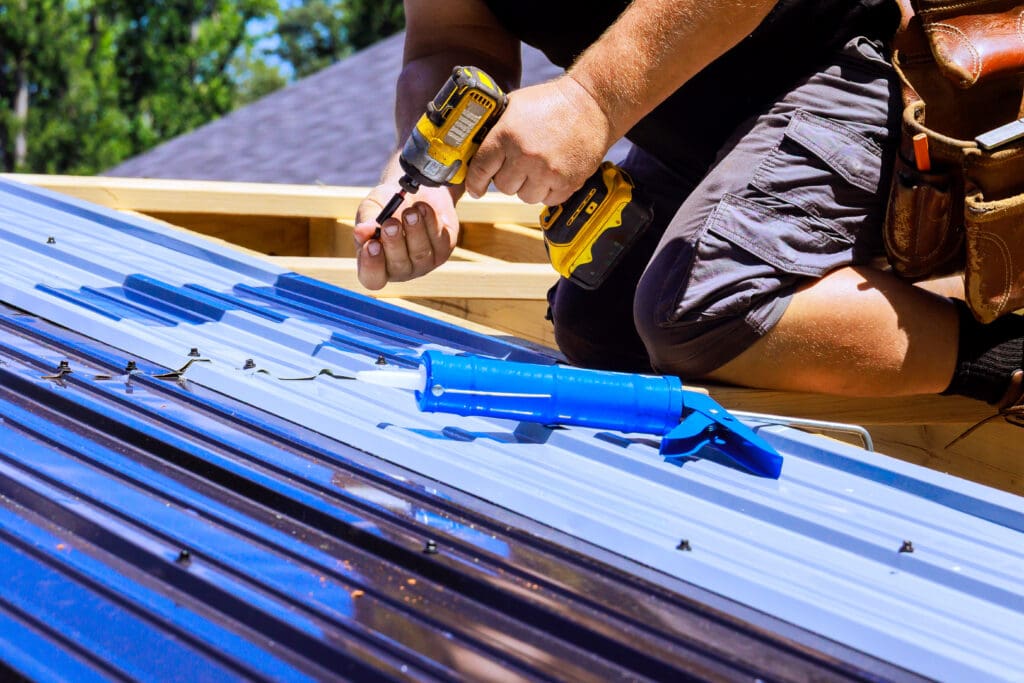 Worker uses drill, sealant while installing metal roof at house.