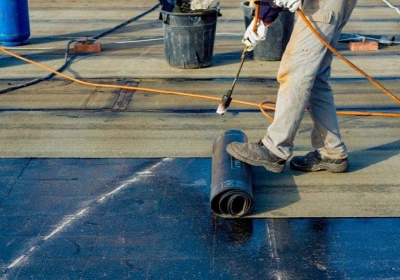 Worker installing rolled roofing membrane with a torch on a flat roof, applying heat to seal seams
