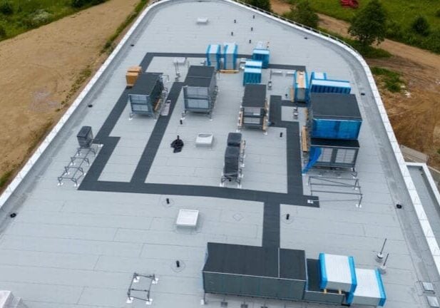 Aerial view of a commercial building rooftop featuring various HVAC units, ventilation systems. The flat roof is mostly gray with black pathways.