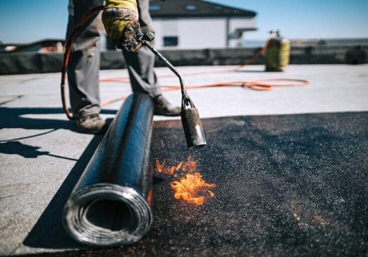 Professional workers insulating rooftop with bitumen membrane. Waterproofing details at construction site. Bituminous membrane waterproofing system details and installation on flat rooftop.